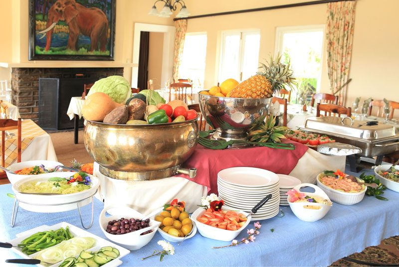 A buffet spread of fruit and vegetables below an elephant painting and a fireplace